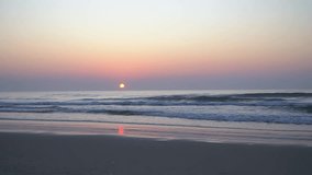 Video of sunrise landscape on sandy beach with waves and rising sun over mist, cloudless sky. And accompanied by the sound of waves and wind.