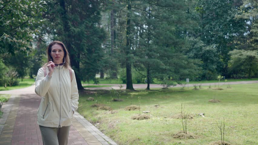 charming adult woman walking in garden with high pines, resting in natural park in spring