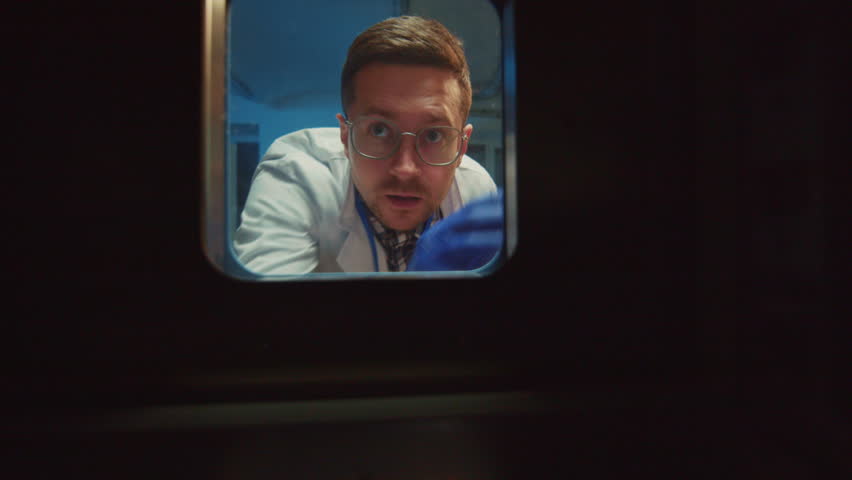 Young attractive medical worker looking into fridge at work. Male research in white lab coat opening door of refrigerator in laboratory in evening. Scientist