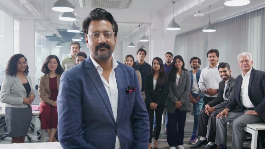 A middle aged Indian Asian confident male entrepreneur standing in front with the diverse team or group of employees at the back looking at the camera smiling in a modern corporate start up office | Shutterstock HD Video #1104433629