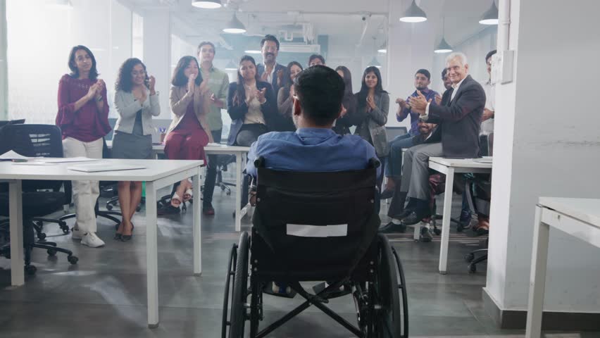 A work achievement of an Indian Asian male handicapped or physically disabled man sitting in a wheelchair cheered by a group of happy office colleagues or team members in a modern corporate start up | Shutterstock HD Video #1104433637