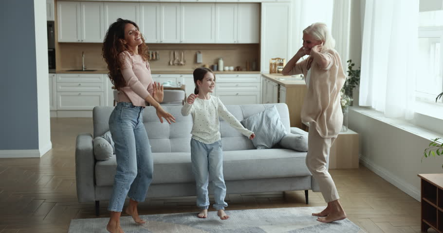 Overjoyed energetic three women generations dance jump barefoot on warm wooden floor in modern living room, listen moving to music, looks carefree, enjoying weekend day, active family have fun at home