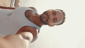 VERTICAL VIDEO: Close up, young smiling bearded male fit athlete uses cell phone, takes selfie while resting after training on the embankment.Backlight