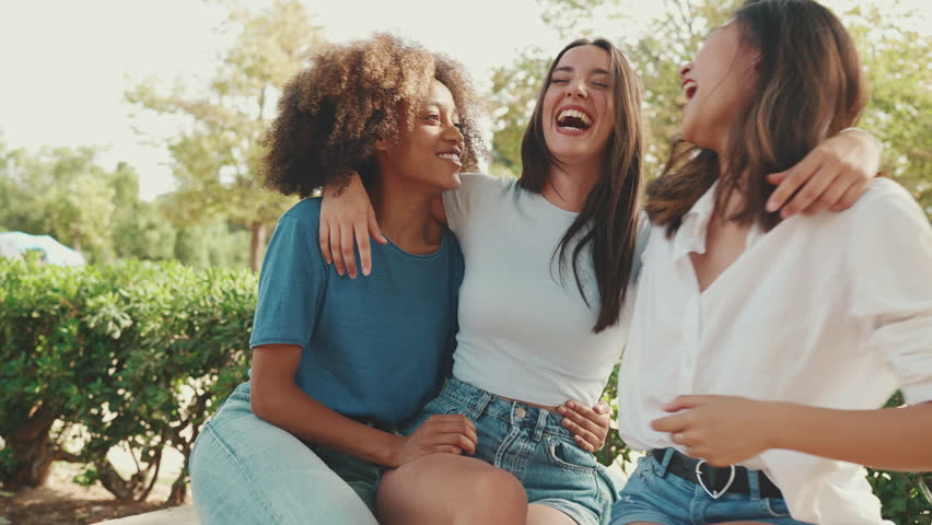 Happy multiethnic young women talking while sitting on park bench on summer day outdoors. Group of girls talking and laughing merrily in city park