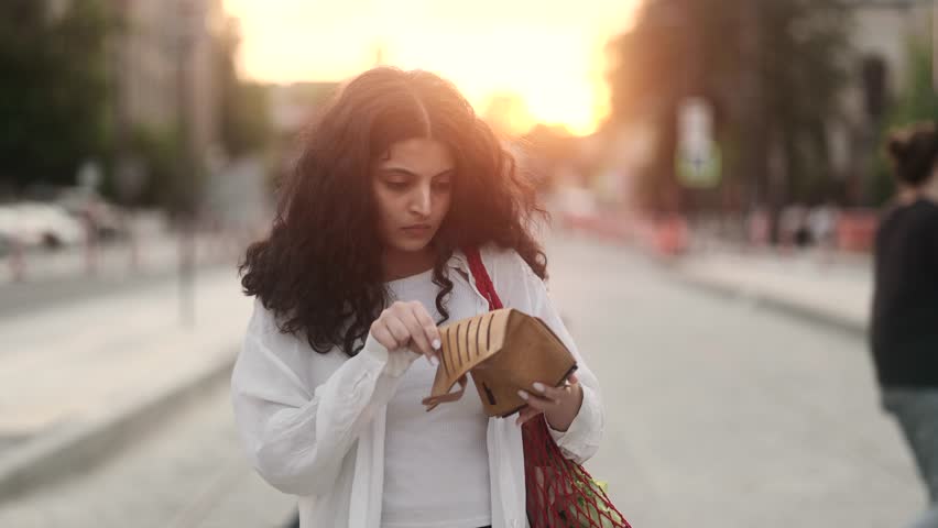 Sad young curly indian woman with shopping eco bag looking at empty purse on sunset street outdoors Upset female with purchases haven’t money cash after shopping and looking at camera shrugs at city Royalty-Free Stock Footage #1104450693