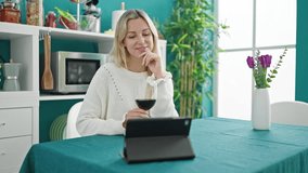 Young blonde woman drinking glass of wine watching movie on touchpad at dinning room