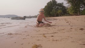 A video moving towards a little boy wearing a hat and swim trunks, digging up the sand on a beach in Thailand during daytime
