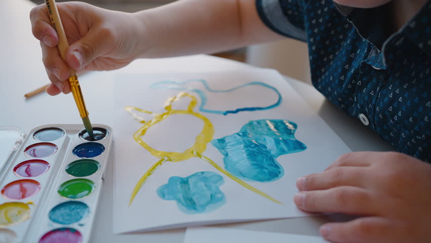 Primary age boy squeezing brush in watercolor paints, drawing picture on paper sheet during art class. Social integration. Education. Children with disabilities. Down syndrome awareness campaign Royalty-Free Stock Footage #1104459283