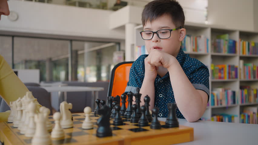 Cute smart child boy with Down syndrome, moves chess pawns on chessboard, during chess battle with his teacher in the classroom. Developing strategic and thinking skills of smart children | Shutterstock HD Video #1104459293