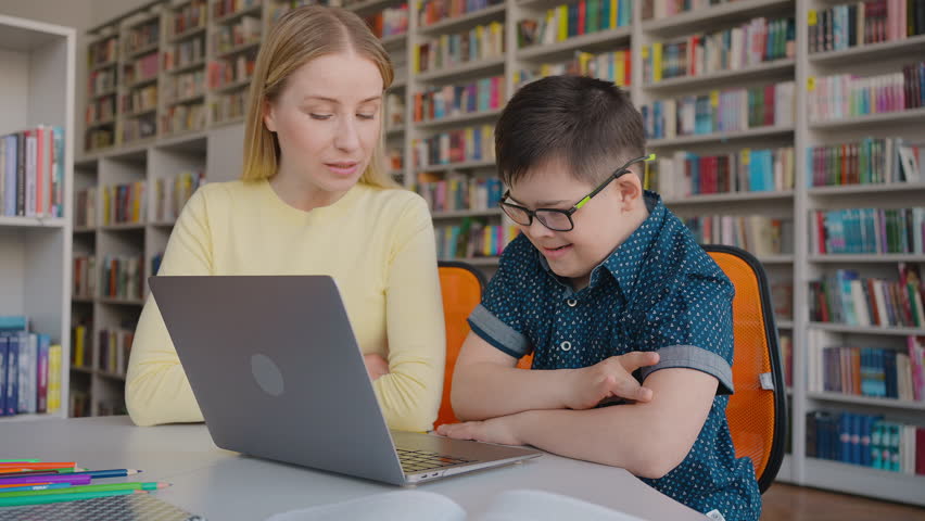 Adorable smart school child boy with disabilities, wearing eyeglasses, sitting at desk, typing text on laptop keyboard, having a lesson with his teacher, educator in the school library campus Royalty-Free Stock Footage #1104459303