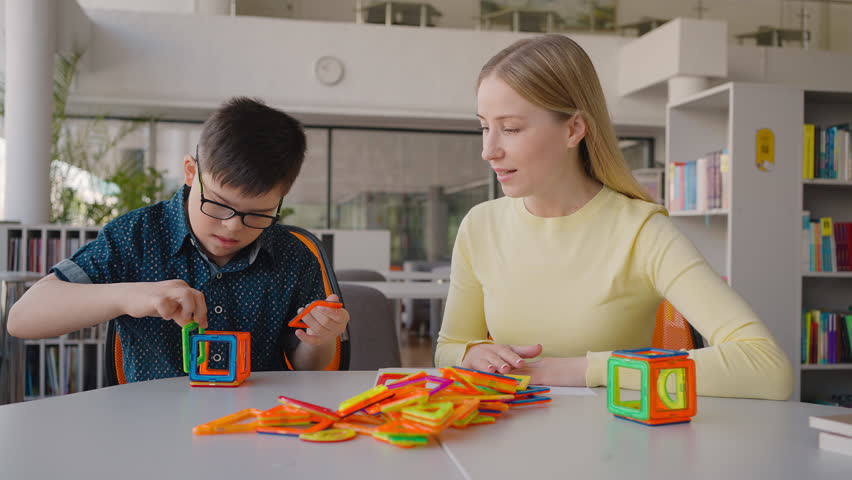 Integration of children with special needs in everyday social and school life. Child boy with Down syndrome plays magnetic constructor with his educational psychologist - teacher in the classroom