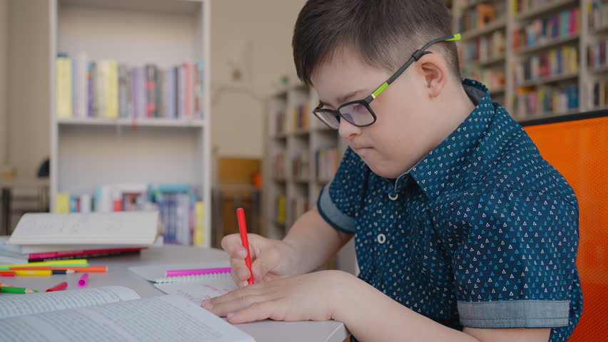 Primary school boy wearing eyeglasses, sitting at desk in the library campus, writing on copybook. Concept of education and social intergration of children with disabilities. Awareness Campaign Royalty-Free Stock Footage #1104459311