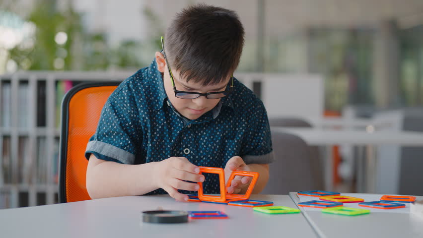Slow motion adorable child boy with Down syndrome, building cube with colorful blocks of a magnetic constructor toy, sitting at desk in the classroom. Educational concept. | Shutterstock HD Video #1104459329