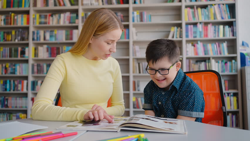 Inspired teacher, tutor, educator engaged in special educational process with Down syndrome schoolboy, sitting at desk in the library campus. The concept of education for children with disabilities.