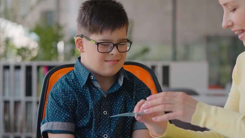 Close-up portrait of a boy with Down syndrome, touching cards during psychological educational class with his experienced teacher. Time to learn. Education and development of elemenatry age kids | Shutterstock HD Video #1104459349