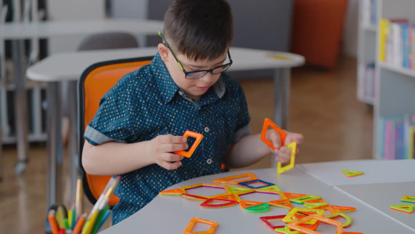 Elementary-aged schoolboy with Down Syndrome plays with colorful blocks of a magnetic construction toy while sitting at a desk in a classroom. Development of motor skills of elementary age kids | Shutterstock HD Video #1104459351