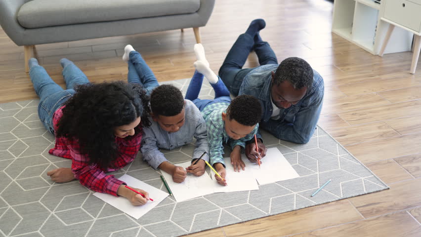 Top view of talented multiracial couple coloring pictures together with focused children while staying at home. Thoughtful parents developing artistic skills of little sons after walk outdoors.