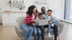 Affectionate african american mom tickling one of cute sons while happy father showing video to another boy. Cheerful parents and children using wireless laptop while having fun at home.