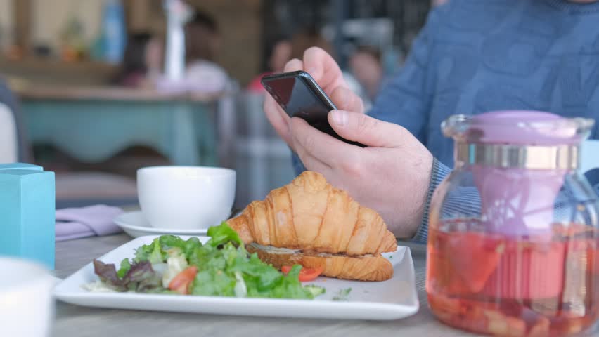 Unrecognizable man businessman sitting at table in cafe cafeteria restaurant with a smartphone in hand prepare eat croissant with salmon and poached egg | Shutterstock HD Video #1104467225