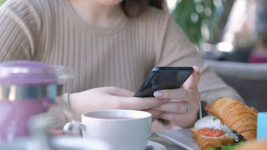 Unrecognizable businesswoman sitting at table in cafe cafeteria restaurant with a smartphone in hand prepare eat croissant with salmon and poached egg | Shutterstock HD Video #1104467229