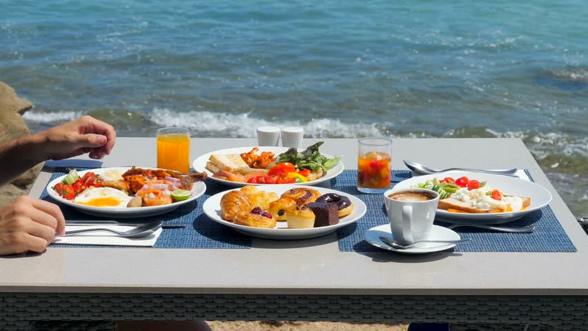 Enjoy a delicious breakfast at a tropical hotel resort. Man drinking orange juice on buffet breakfast at a luxurious beachfront villa. Dining at a beachside restaurant | Shutterstock HD Video #1104468359