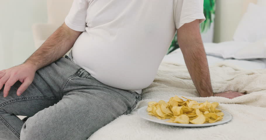 unrecognizable boy touching his belly fat sitting on the bed near potato chips. Royalty-Free Stock Footage #1104470411