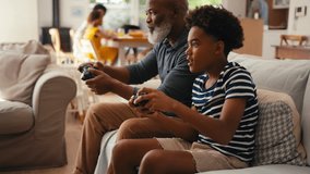 Grandfather and grandson sitting on sofa at home holding controllers playing video game together with multi-generation family in background - shot in slow motion