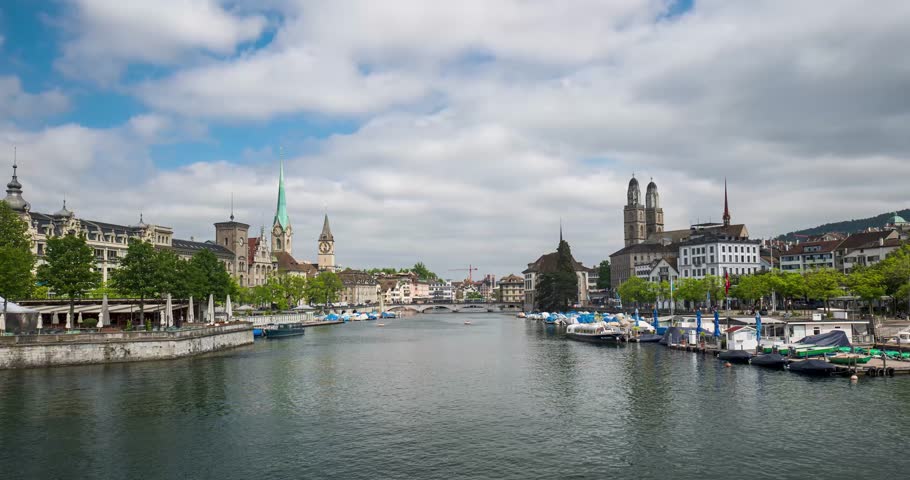 Zurich time lapse from Quai bridge towards Munster bridge. Wide-angle view of Limmat river, Grossmunster and Fraumunster churches, parked sailboats, pedestrian and cable car traffic. Moving clouds, sp