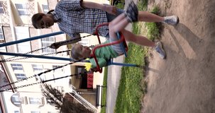 Vertical video. A young girl in stylish shorts and a plaid shirt is riding a small two year old child on a swing. The child enjoys holding onto the swing with her hands.