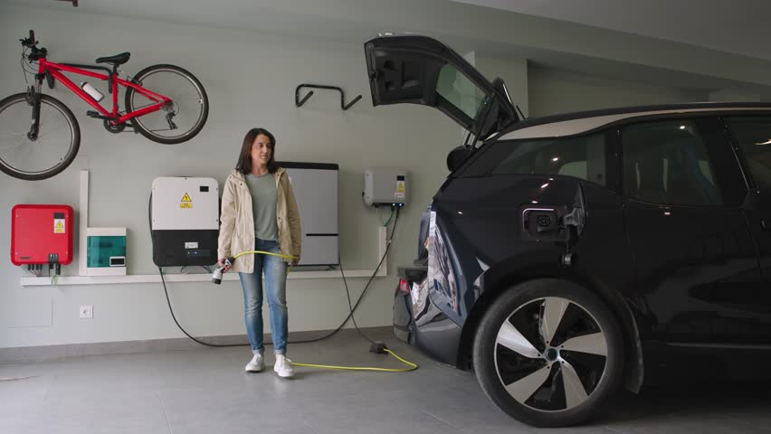 Woman comes home, plugs a charger into an electric car socket in the garage, and pulls a bag of groceries out of the trunk. Sustainability, Energy Efficient Domestic Life. Horizontal 4k video. Royalty-Free Stock Footage #1104482307
