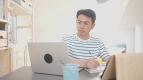 Video of an asian adult man writing notes while looking the screen of the laptop in a home office