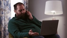 A man in a green sweater sits in a chair and talks on the phone. A bearded male freelancer sits in a chair with a laptop on his lap and discusses a new work project with his employer on the phone.