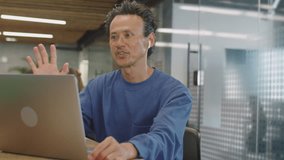 Medium shot of Asian man in wireless earphones speaking via video call on laptop during day in coworking room