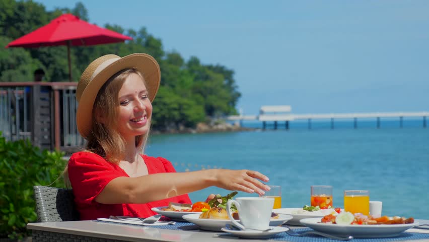 Young woman sitting at table with plates full of food in restaurant with sea view. Girl on buffet breakfast in luxury resort. Travel lifestyle. Summer vacation. | Shutterstock HD Video #1104490845