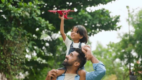 Happy smiling indian girl kid playing with airplane toy by sitting on father shoulder at park - concept of freedom, family support and togetherness : vidéo de stock