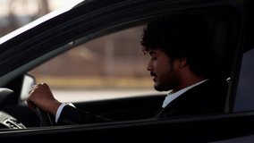 A young Indian businessman sits in a car and is nervous about being stuck in a traffic jam. A curly-haired man in a business suit is talking on the phone while sitting in a car.