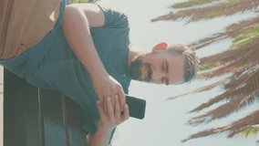 VERTICAL VIDEO, Young smiling man with beard sits on bench, uses cellphone. Backlight