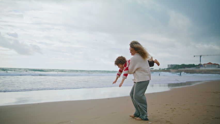 Joyful mom spinning kid on autumn beach. Smiling babysitter playing airplane holding curly boy in hands. Beautiful young mother playful son having fun together at seaside. Loving family relationships Royalty-Free Stock Footage #1104501665