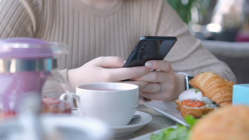 Unrecognizable businesswoman sitting at table in cafe cafeteria restaurant with a smartphone in hand prepare eat croissant with salmon and poached egg | Shutterstock HD Video #1104505027