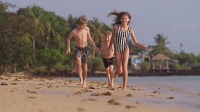 A daytime video of three kids holding hands while happily running in slow motion along the shore of a beach in Thailand