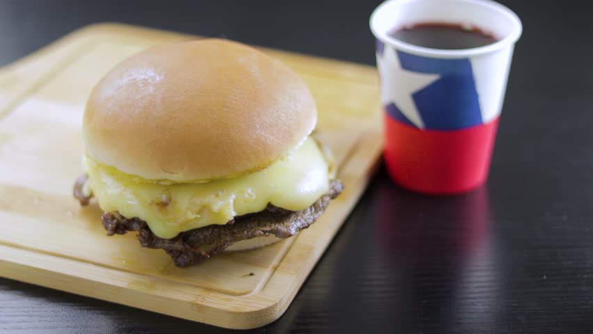 
Barros Luco sandwich popular in Chile: meat with melted cheese, hand taking paper cup with Fiestas Patrias wine, on black wooden table. with copy space concept of typical Chilean food