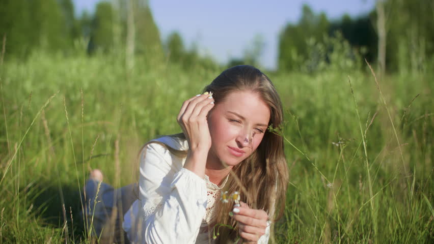 Attractive young happy smiling woman lying on field with chamomile flowers in hands. Portrait of pleasant female enjoying resting relaxing on nature tearing petals from flower in summer sunny day.
