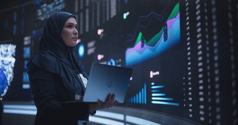 Muslim Female Data Center IT Engineer Standing in a Room with an AI Neural Network Settings on a Digital Screen. Cloud Computing Expert Uses Laptop for Servicing the System in Cyber Security Facilityの動画素材