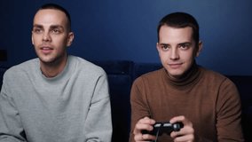 Smiling friends with gamepads joystick controller playing video game on sofa at home. Portrait of caucasian guys playing game with console relaxing on couch at home.