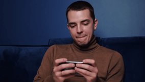 Close up caucasian man playing game on phone and looking concentrated and happy. Handsome man holding phone horizontally and using it while lying in bed at home.