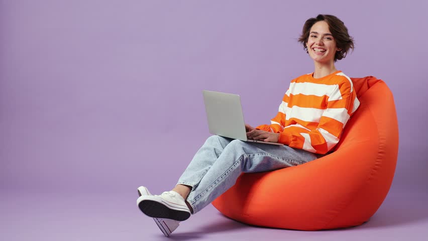 Full body caucasian young student fun smart IT woman wear casual clothes sit in bag chair hold use work on laptop pc computer isolated on plain pastel light purple background. People lifestyle concept
