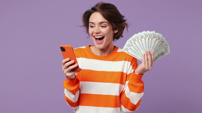 Excited happy joyful young woman wears casual clothes use mobile cell phone hold win fan of cash money in dollar banknotes isolated on plain pastel light purple background. People lifestyle concept