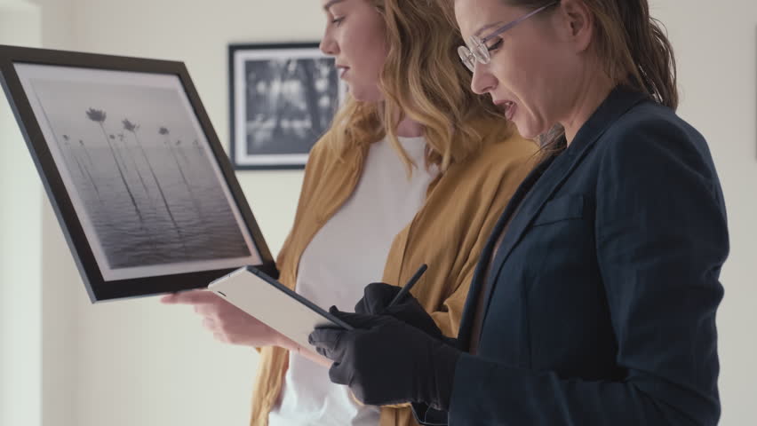 Medium shot of female artist discussing framed photo with gallery curator while she using digital tablet in exhibition hall