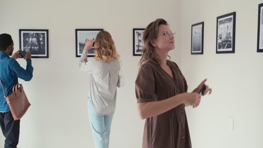 Medium long shot of woman watching photo exhibition in art gallery and taking pictures with mobile phone, other visitors walking behind