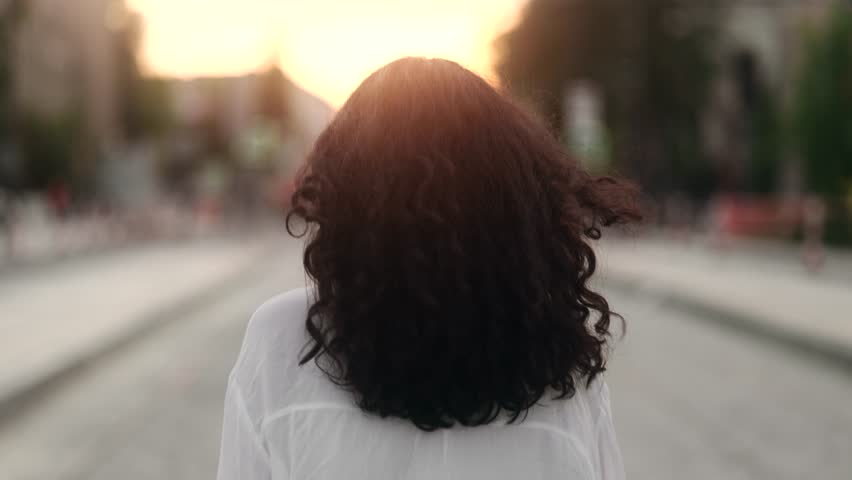 Charming young indian woman walking down the street turns around with flying hair and looking at camera outdoors Happy relaxed lady walking on the city centre enjoying beautiful sunset alone